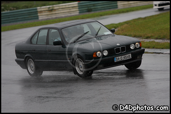 E34 525i - Page 1 - Readers' Cars - PistonHeads - The image portrays a black BMW driving on a wet race track with a heavy mist. The rain-soaked car is captured mid-motion, leaving behind a trail of spray on the wet asphalt. The track is illuminated by ambient lighting, emphasizing its surface and the car's motion. The focus of the photo is the car, its design and the droplets captured in its wake emphasizing its speed and movement.