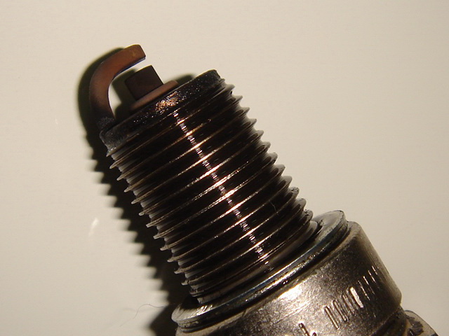 This image features an object with a metallic base and a protruding spindle, suggesting it might be a mechanical component or a tool partially wrapped in black tape. The spindle itself has several layers with ridges, giving it a cylindrical and hexagonal shape which could be indicative of a screw-like function. It is positioned against a plain white background, emphasizing the object's structure and texture. The black tape appears to be coiled, possibly serving a purpose in the object's intended use such as for insulation or securing parts. The object's overall design and appearance suggest it may be part of an engine, a manufacturing device, or a piece of industrial equipment.