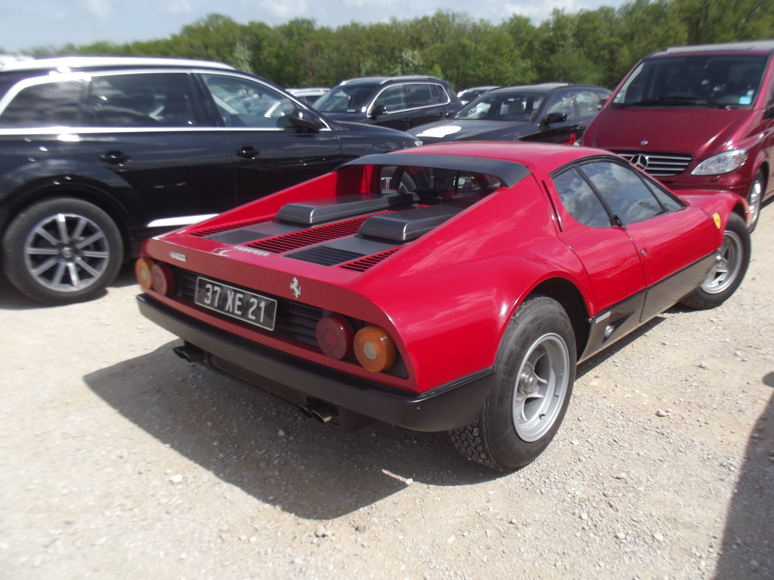 COOL CLASSIC CAR SPOTTERS POST!!! Vol 2 - Page 397 - Classic Cars and Yesterday's Heroes - PistonHeads - The image showcases a vibrant red Ferrari car parked on a gravel lot. The car is sleek and well-maintained, featuring a black convertible top and a set of shiny silver alloy wheels. In the background, a line of black cars can be seen parked in a neat row, adding contrast to the vivid red of the Ferrari. The lot is nestled between trees, offering a slightly blurred backdrop to the scene. The overall setting suggests a private display or showroom for exquisite vehicles.