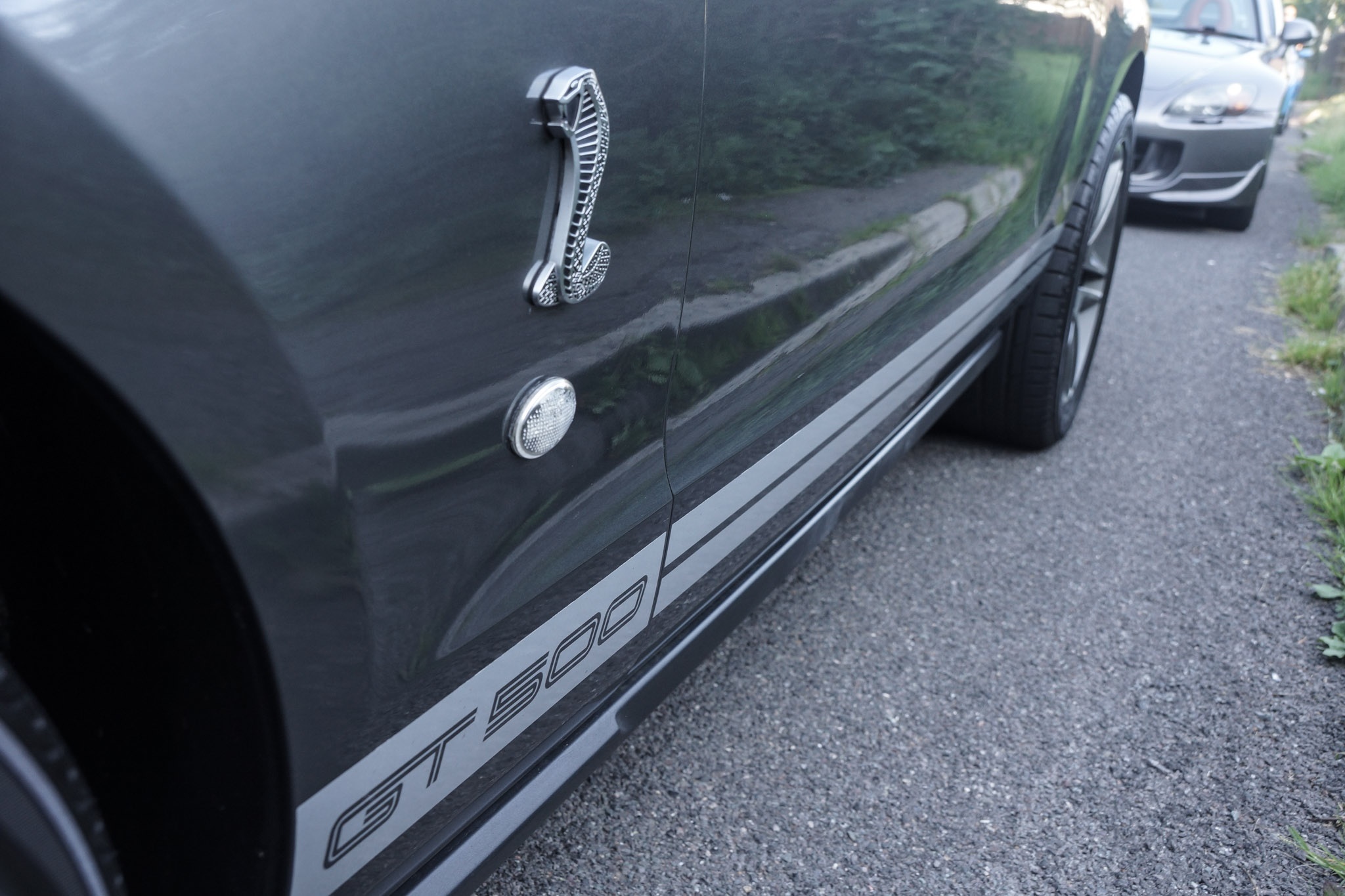 Bristol Tunnel Run 2015 - Page 5 - South West - PistonHeads - The image shows a close-up view of a black car, likely a sedan, with a shiny metallic door handle. The car is parked on a weathered gray gravel or pavement surface that appears worn, perhaps from frequent use. Reflections can be seen on the car's sleek body, which suggests a well-polished and possibly recently cleaned surface. The daylight indicates a clear and sunny day.