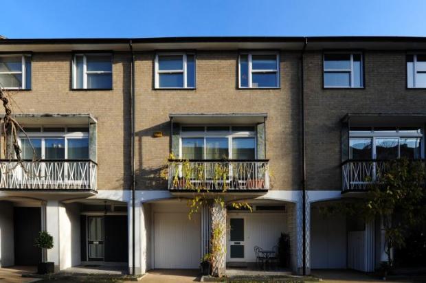 A building that has a clock on the side of it - Pistonheads - The image depicts a row of multi-story buildings, each with a balcony on the second floor and large windows. The architecture of the buildings suggests a European or American style, with concrete construction. The balconies have a simple white railing and each is furnished with plants, adding a touch of green to the otherwise neutral color scheme of the buildings. A staircase leads to the upper levels, indicating the buildings have more than one entrance. The sky is clear, suggesting it is a bright and sunny day.