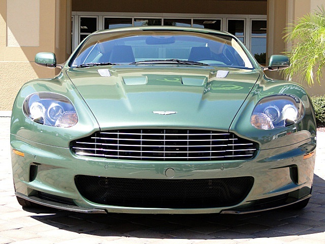 Damn that Tony Hall !! - Page 1 - Aston Martin - PistonHeads - The image features a distinctive green Aston Martin sports car parked on a brick pavement. The car's sleek design and the prominence of the badge on the grille are clearly visible. The front of the car is framed by a thin strip of metal, adding to its sporty aesthetic. The vehicle takes up the majority of the frame, demonstrating its impressive size and presence. The background is relatively nondescript, with only a building facade providing context for the location. The focus of the image is clearly on the car, which is the main subject.