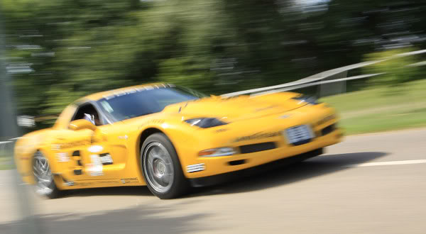 Ish Pistonheads Beast Night Yellow - The image shows a yellow sports car in motion on a road. It appears to be a blurry action shot, possibly of a pro driver or a racing car, considering the intensity of the scene. The car has a sleek design with a curved front hood, and there are numbers, which could indicate a racing context or a specific model. The surrounding environment is forested, suggesting the road is set amidst trees, adding a natural backdrop to the shot.