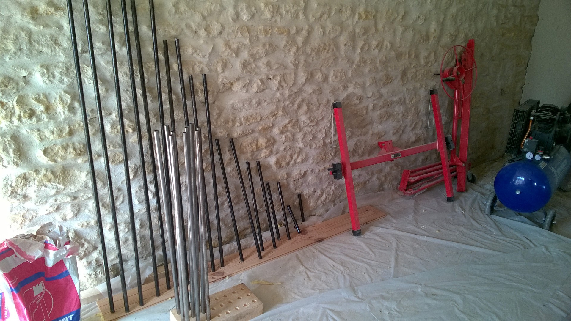 Our French farmhouse build thread. - Page 13 - Homes, Gardens and DIY - PistonHeads - The image shows an indoor scene where several metal poles are arranged on a structure, possibly on a construction site or in a manufacturing area. The poles appear to be metal pipes or rods, and they are held in place by wooden supports and clamps. A red made-like scaffolding or frame is nearby, suggesting some degree of setup or construction activity. The background features a brick wall, and there's some industrial equipment or machinery in the corner of the frame. The atmosphere suggests a practical, utilitarian space.