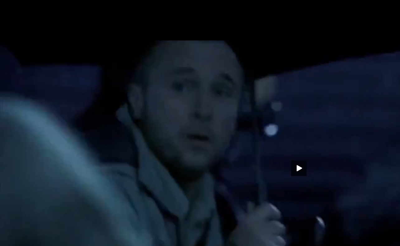 A man in a suit and tie sitting in a car - Pistonheads - This image appears to be a still frame from a video. A man is seen in the foreground, from the chest up, holding what looks like a light source under a car hood. The background is blurry but suggests an open area with a dark sky at dusk or night. The man seems focused on the task at hand, possibly working on or under the car. There is a play icon on the image, indicating that this may be a screenshot from a video streaming service.