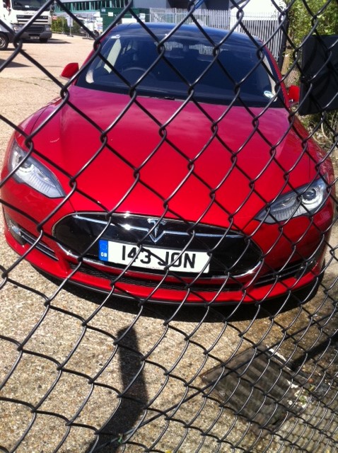 Tesla S spotted, RHD but a May 14 car? - Page 1 - EV and Alternative Fuels - PistonHeads - The image shows a striking red Tesla Model S car parked on a paved surface. The vehicle is positioned at an angle with the front driver's side facing towards the left side of the image. It has a sleek and modern design, with a clean and uncluttered appearance. The license plate starting with "143" is prominently displayed on the front bumper. The car is enclosed within a chain-link fence of a parking lot, which adds a sense of security or containment to the scene. The fence's texture and structure create a visual mesh that partly obscures the car while still allowing its vibrant color to stand out.