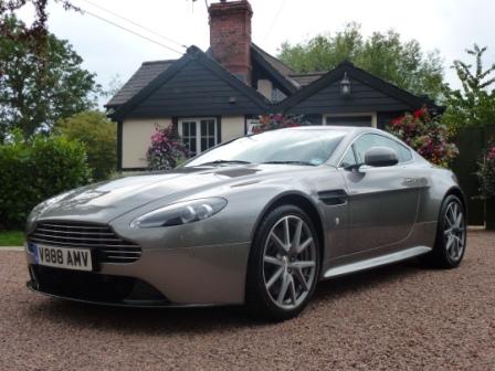 What is a Vantage S? - Page 2 - Aston Martin - PistonHeads - This image features a silver Aston Martin sports car parked on a driveway in front of a house. The car is sleek and shiny, standing out against the natural tones of the driveway. It has black wheels and a black soft top that contrasts with the silver body. The house behind the car is a two-story structure with a shingled roof, surrounded by lush green trees. The sky is overcast, suggesting an overcast or possibly rainy day.