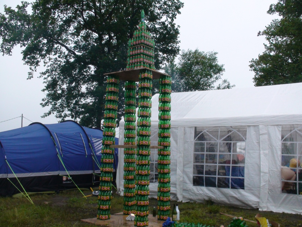 Beer Mountain in style  - Page 1 - Le Mans - PistonHeads - The image shows a unique outdoor sculpture in a grassy area, composed of multiple cans arranged in a pyramid-like structure, with the largest element at the top. Surrounding the sculpture is a pristine white tent, and partially visible tents of a contrasting blue hue. Some people are present in the background, observing the artwork. The sky overhead is overcast, suggesting a possibly cool or cloudy day.