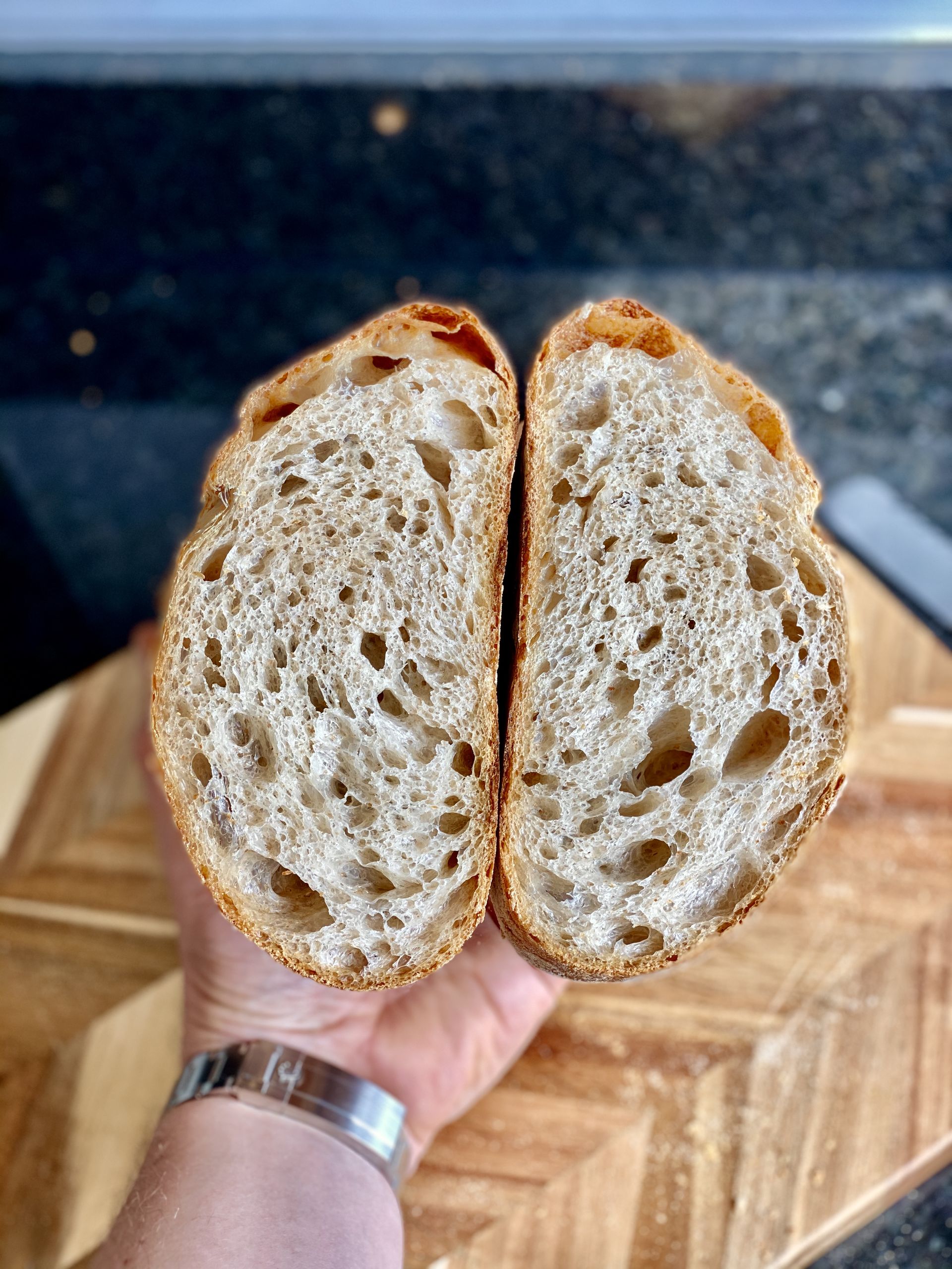 Sourdough breadmaking - Page 17 - Food, Drink & Restaurants - PistonHeads - The image features a large, freshly baked loaf of bread placed on a cutting board. The bread is sliced in half, revealing the soft interior with visible crumb structure. It's being held by a person whose hand appears at the bottom of the frame. The background is blurred but suggests an indoor setting, possibly a kitchen or bakery. There's no text present in the image.