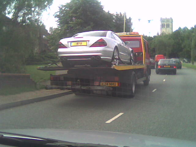 Pistonheads Lambo Spotted Mph - The image captures a scene on a street where a silver sports car is on a tow truck. The car is positioned in the back of the tow truck and secured by chains, indicating it might be inoperable or moved for some reason. Two people are visible in the image, possibly the tow truck driver and the owner of the car. There are other vehicles present in the image, a red van and a black car, which suggests it's a busy street. The perspective of the photo suggests it might have been taken from the front seat of a vehicle in front of the tow truck.