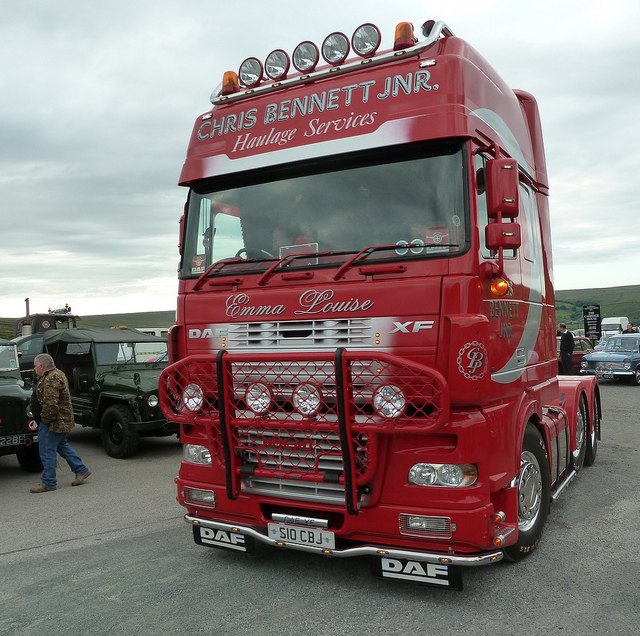 Scania 141 Astrans for wedding transport - Page 1 - Commercial Break - PistonHeads - In the image, a large red truck with a cab and a trailer is parked on a road next to a green jeep. The truck is adorned with the sign "Chris Bennett JNR." The jeep's distinctive green color stands in contrast to the red of the truck. A man is standing on the right side of the image, looking towards the truck. The setting appears to be an open lot or a similar outdoor space. The horizon in the background suggests that the location is relatively open or consists of a wide road.