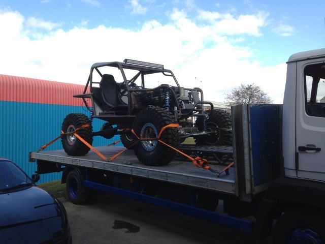 Road legal Off road buggies - Page 1 - Off Road - PistonHeads - In the heart of the image, a robust off-road buggy commands attention. It's positioned on a sturdy flatbed trailer that contrasts sharply with its white and blue cab, suggesting a recent haul. The buggy itself is a blend of black and gray, a testament to ruggedness and durability. Orange wheels add a pop of color, hinting at this off-road vehicle's readiness for exploration. The backdrop is a serene blue sky, dotted with fluffy white clouds, creating a peaceful contrast to the adventurous spirit embodied by the buggy.