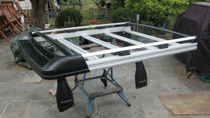 Roof Rack Pistonheads - The image shows a large window hinged to a wooden frame, which is old and worn. The window is leaning on a blue wooden cart, possibly for storage or moving. The setting seems to be an outdoor space with a background that includes a wooden fence, potted plants, and a lone chair. The focus of the image is the window, which is placed in such a way that it leans backwards, creating an interesting diagonal effect. The colors in the image are mainly the shades of brown from the wooden elements and the contrast provided by the blue cart.