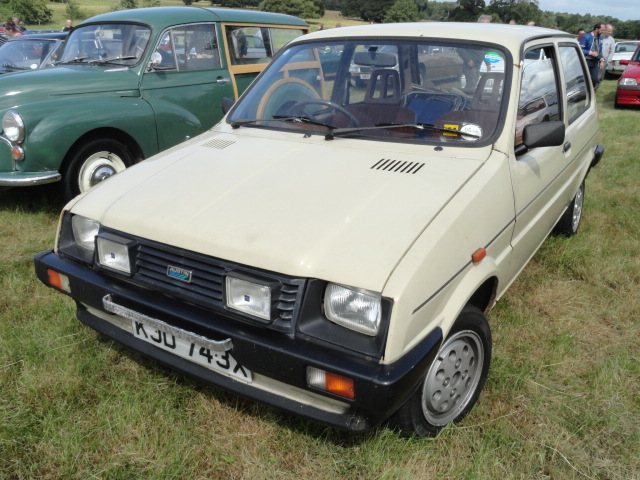 RE: Festival of the Unexceptional | PH Gallery - Page 7 - General Gassing - PistonHeads - The image shows a vintage car displayed at what appears to be an outdoor show or exhibition. The car is beige and has a noticeable sunroof, with the roof down. It features a spare tire mounted on the back door, and there are visible license plates and a registration plate. The vehicle's design suggests it could be from the 1980s to 1990s. In the background, there is an open field where other vintage vehicles can be seen, indicating that this car is part of a larger display. There are no people visible in the image.