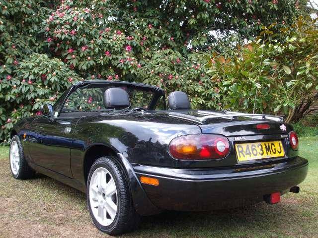 Ive Pistonheads Elite Joined - This image shows a black sports car parked in what appears to be a grassy area, surrounded by trees with pink flowers. The car is a two-door convertible with a black soft top and black hard top. It is equipped with a black spoiler. The license plate of the car reads R4463 MGJ, indicating it may be a Mazda Miata, a popular model for sports car enthusiasts. The setting suggests a serene, natural environment, perhaps a park or a garden.