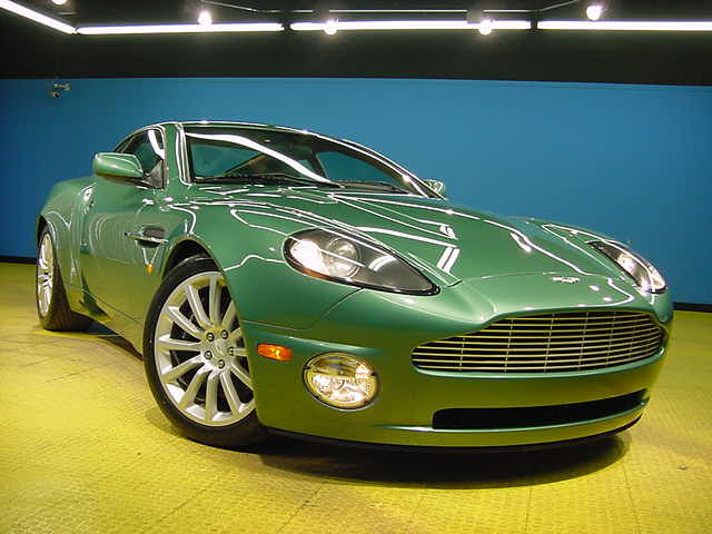 Green Vantage Pistonheads - The image shows a gleaming green two-door sports car parked indoors, likely in a showroom. The vehicle is new and appears in excellent condition, with a striking green paint job that emphasizes its aerodynamic design. The front grille and badge are prominently visible, and the wheels and engine vents are also clean and reflective. The car is displayed against a contrasting blue background, which adds to the dramatic presentation. The interior lighting provides a soft, diffused illumination, enhancing the car's glossy and polished appearance.