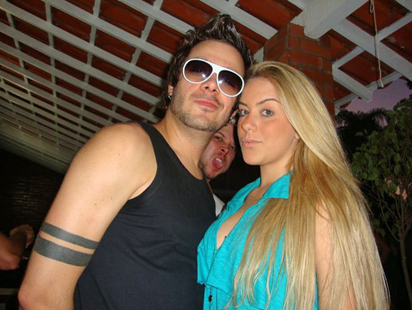 Bbb9 Max - The image depicts a man and a woman posing closely together. Both individuals are wearing sunglasses and have tattoos on their left arms. The woman has blonde hair and is wearing a light blue top. The man has dark hair and is dressed in a black sleeveless top. They appear to be at a social event, as there's a structure with tiles visible in the background.