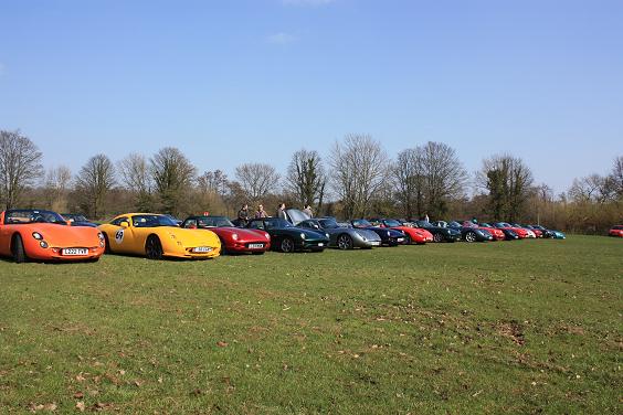 Valley Cobweb Tvrcc Pistonheads Tees - The image shows an outdoor parking area filled with unique sports cars, exuding a vibrant gathering. The vehicles feature branding and vibrant colors, suggesting they are likely on display or participating in a car show. The setting appears to be a grassy field with open visibility, allowing for a dynamic and immersive view of the vehicles. In the background, there are trees suggesting a natural environment, and the vast sky above adds an overall open and calming atmosphere to the scene.