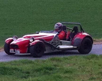 A red and white motorcycle parked in a field - Pistonheads - This image captures a single-seat, red and black racing car in motion on a roadway. The car, designed with two wheels in the front and two in the back, is resembling the cars raced at Le Mans. The driver, dressed in a full racing suit, seems to be skillfully maneuvering the vehicle, which also features a protective windshield. The setting appears to be a rural area, with a grassy verge visible on the side.