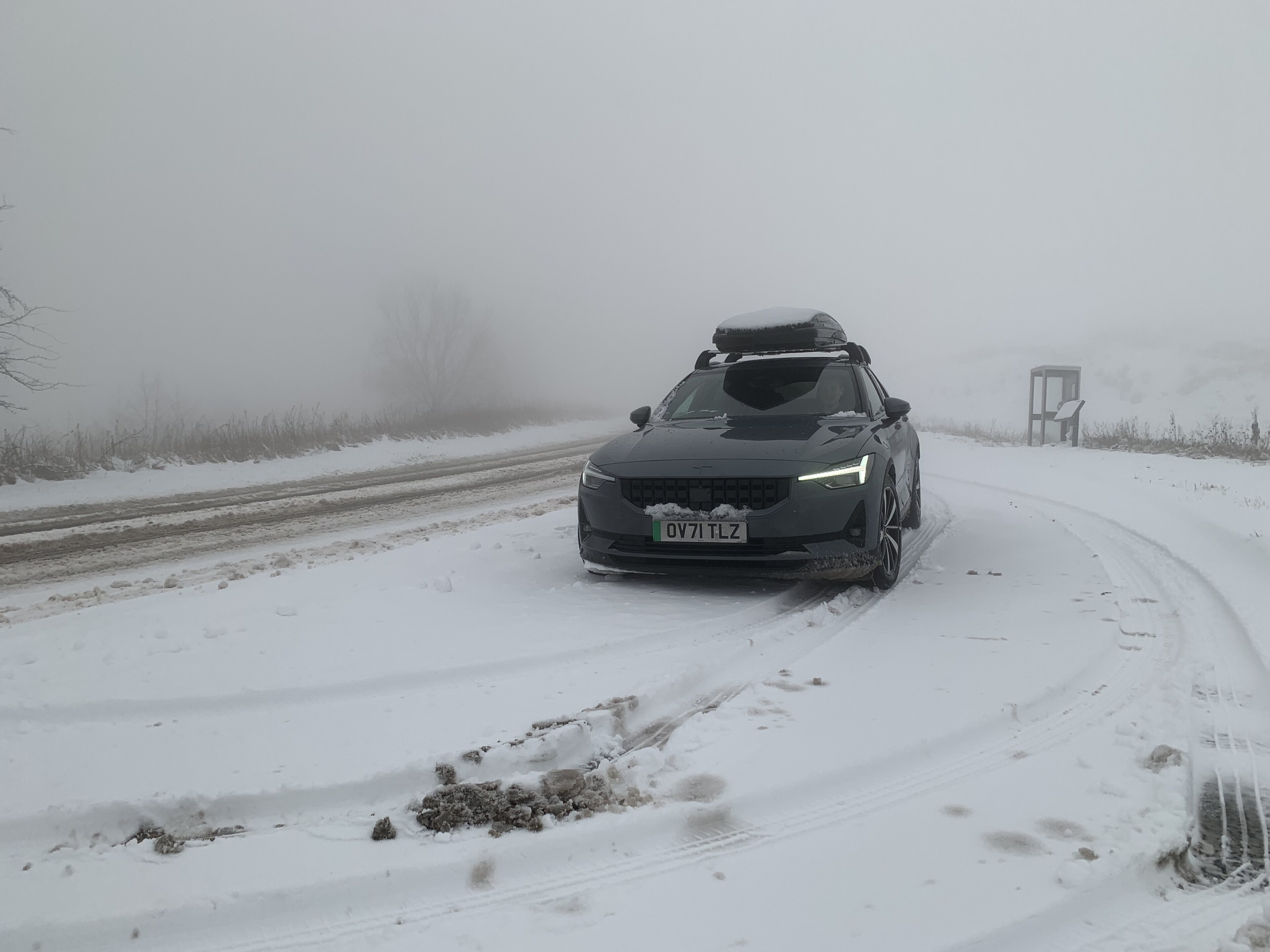 EVs... no one wants them! - Page 470 - Car Buying - PistonHeads UK - The image shows a snowy scene with a car parked on the side of a road. The car is partially covered in snow, indicating recent or ongoing snowfall. There are tracks in the snow around the vehicle, suggesting it has moved recently. On top of the car's roof rack is a set of skis and poles, hinting at winter sports activity in this area. The sky above appears overcast, contributing to the wintry atmosphere. A road sign is visible on the left side of the image, but the content of the sign is not clear due to the resolution.