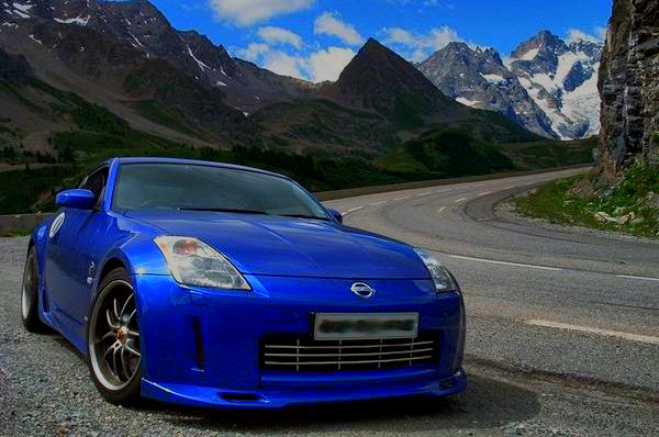 Some half decent pics - Page 1 - North West - PistonHeads - The image shows a blue Nissan sports car parked on the side of a winding mountain road. The car, which is the main subject of the image, is positioned in the bottom right corner. The surrounding environment features majestic mountain peaks and a clear blue sky above. The road curves gently around the scene, leading the eye towards the vast wilderness in the background. The contrast between the sleek modernity of the car and the rugged natural beauty of the landscape creates a striking visual.