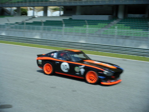 Pistonheads Afos Sepang - This image depicts a race car on a track. The car is painted in a striking orange with black and white accents, indicating it's possibly for competitive racing. Its design suggests it's an open-wheel sports car, a common type in motorsports. The track appears well-maintained with green grass lining the sides, and there are empty green stands surrounding the track, implying the photo might have been taken before an event or during a practice session. The photo itself appears to be a still from a high-speed video or a timelapse, as indicated by the motion blur on the edges of the image.