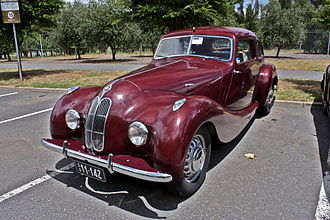 Pistonheads - The image is in color and displays a shiny maroon vintage car parked in a paved lot. The car features the distinctive, vintage styling typical for vehicles of this era, with rounded bodywork, a prominent grille, and flared wheel arches. 

The vehicle is well-maintained, with a glossy finish on its exterior that reflects the surrounding environment. The lot itself is adjacent to what appears to be a row of trees, suggesting the setting might be a park or a car show. 

The image does not contain any visible text or branding. The focus is solely on the car, which occupies a central position within the frame, drawing the viewer's attention immediately. The car is oriented toward the right side of the image, possibly towards an exit or a main road.