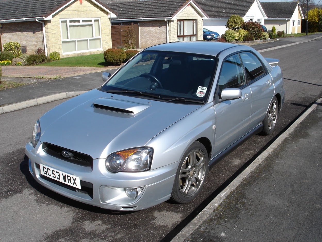 RE: Shed of the Week: Subaru Impreza WRX PPP - Page 4 - General Gassing - PistonHeads - This image shows a silver-colored car parked on the side of a road. The car is a hatchback with a visible spoiler and what appears to be a sporty, modified design. It has four doors and a black roof rack. The vehicle is parked next to a sidewalk and is partially covered by the shadow of trees, suggesting it might be a residential area. In the background, there are houses lining both sides of the street.