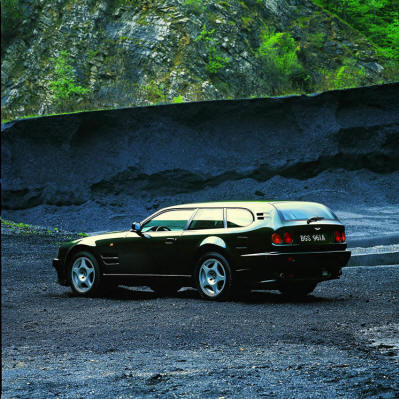 Pistonheads - The image features a sleek, dark-colored car with shiny accents, parked on a gravel road. The car is positioned in the center of the image, facing towards the camera. The road is flanked by a steep hill on one side and a verdant, lush hill on the other, creating a contrast between the urban vehicle and the natural landscape. The sky appears to be overcast, casting a soft, diffused light over the scene. The overall mood of the image is mysterious and somewhat moody.