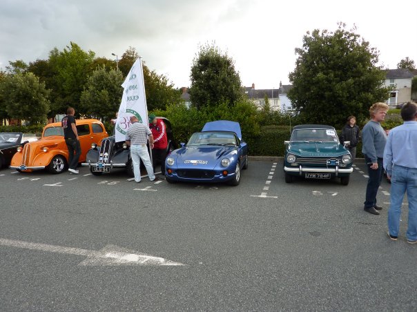 September Pistonheads Pembrokeshire County - This image showcases a convivial gathering of people in a parking lot. There are multiple people milling about, with one individual who stands out standing next to an orange car to the left side of the frame. The cars are parked neatly in rows, creating a grid of vintage vehicles. Notably, one of the cars has a flag attached to its side mirror, adding a touch of color and flair to the scene. The overall setting suggests an outdoor car show or exhibition. The atmosphere appears to be casual and friendly, with attendees enjoying the sight of these classic vehicles.