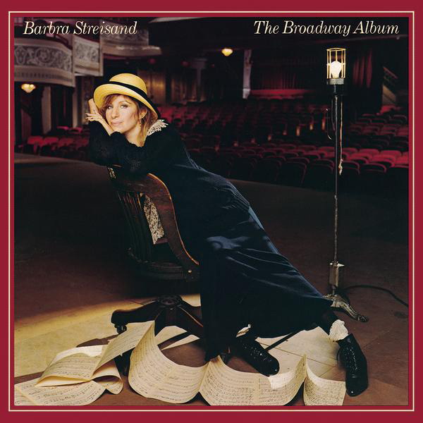 This is an album cover featuring a woman in a theatrical setting. The woman is dressed in what appears to be a style from the mid-20th century, with a long coat and a large hat. She is seated in a red chair and leans against a lamp. She is looking downwards and appears relaxed. The backdrop is a dark theater with rows of red velvet seats leading up to a stage. The album title "Broadway Album" and the artist's name "Barbra Streisand" are prominently displayed at the top of the cover. The overall impression is one of classic elegance and musical theater charm.