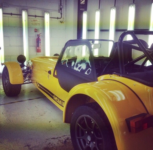 Hello, I've ordered a Supersport 140 - Page 5 - Caterham - PistonHeads - The image shows a bright yellow and black sports car on a car lift. The car has a prominent front grille and a streamlined body, with an open roof, likely a convertible. It's parked inside what appears to be a garage or workshop, and there are fluorescent lights illuminating the interior and shining onto the car. The ambiance is that of a professional, possibly race-related, setting.