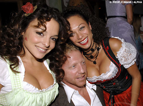 Cryder - The image is a color photograph of three people. At the center, there is a man standing between two women. The woman on the man's left has curly dark hair, wears a green and white dress with puffed sleeves, and appears to be smiling. The woman on the man's right has straight dark hair, wears a black traditional bodice and black pearl necklace, and also seems to be smiling. The man has short, light-colored hair, wears a dark jacket over a white collared shirt, and has a neutral expression on his face. The women's attire suggests a theme or costume party. The background is blurred and indistinct, focusing attention on the three central subjects.