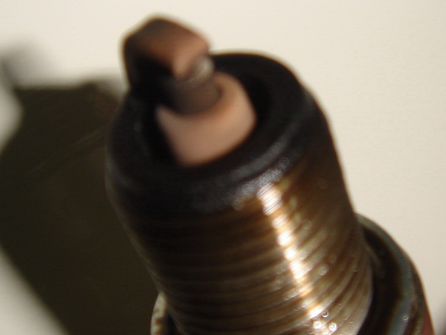 The image features a close-up of a mechanical part, specifically a tapered shaft or spindle. It appears to be made of metal with an oil film visible on the surface. There's a distinctive bronze coloring at the base of the shaft, which might indicate it is coated or has been treated for certain purposes. The background is out of focus, emphasizing the object in the foreground. The style of the image suggests it could be used to illustrate mechanical components or as part of an instructional material related to machinery or engineering.