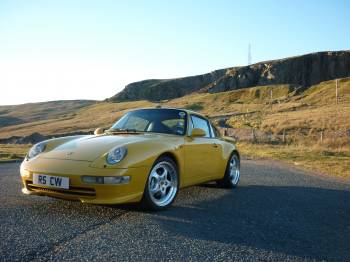 took the car for it's mot today...... - Page 1 - Porsche General - PistonHeads - The image captures a sunny day with a vibrant yellow Porsche parked on the side of a highway. The car is positioned on the pavement, its sleek design accentuated by the sunlight casting long shadows. The backdrop features picturesque scenery with a rocky outcropping and a patch of grassy terrain, suggesting a countryside or park setting. Despite the absence of people, the car is the focal point of the image, signifying it's in use and possibly about to embark on a journey.