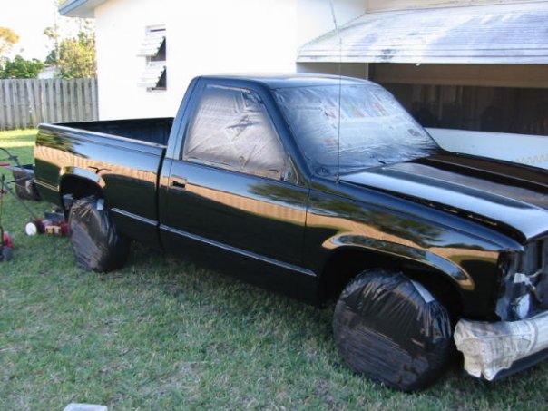 Pistonheads - This image shows a dark-colored pickup truck parked on grass. It appears to be a four-door vehicle with a visible spare tire mounted on the back of the cab. The truck's glass is frosted or tinted, and there are bags or protective covers over the front wheel wells and front bumper. Surrounding the truck, there are parts of what could be gardening or landscaping equipment, suggesting some maintenance or work is happening either on the truck itself or nearby. In the background, there is a partial view of a house with white walls and a wooden fence.