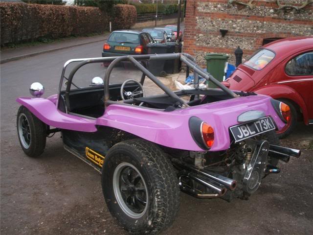 Sun Pistonheads Shine - The image shows a vibrant purple sidecar trailer parked on a gravel surface next to a street. The trailer has a black canopy and a folding windshield. In front of the trailer, there's a wooden fence. Additionally, there are two red insect-themed cars in the background. The grass verge nearby is lush and green, indicating a healthy climate or summertime. The sky is not visible, and the overall atmosphere of the image is cheerful and nostalgic.