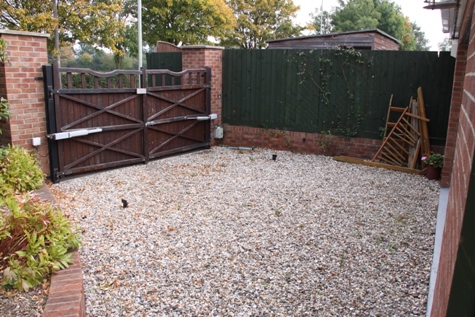 Pistonheads Driveway Small - The image shows a privately owned property with a well-maintained garden. The main feature is a sturdy wooden gate with two large vertical bars and two smaller horizontal bars, closed and secured in its tracks. This gate is mounted within a tidy reddish-brown brick wall that surrounds the property, providing a sense of privacy and security. Beyond the wall, there is a section of green fence, suggesting the presence of an adjacent property next door. On the ground in front of the gate, there appears to be a mixture of gravel, stone, and possibly soil, contributing to the landscaped appearance of the front yard. There is no visible text or distinctive branding in the image. The surrounding landscape is filled with greenery and other plants, which are well-groomed and contributing to the overall natural beauty of the property. The sky is overcast, indicating that the photo was taken on a cloudy day. There are no people or moving vehicles in the scene, and there are no indications of current activity, suggesting the photo may have been taken during a quiet time of day or week.