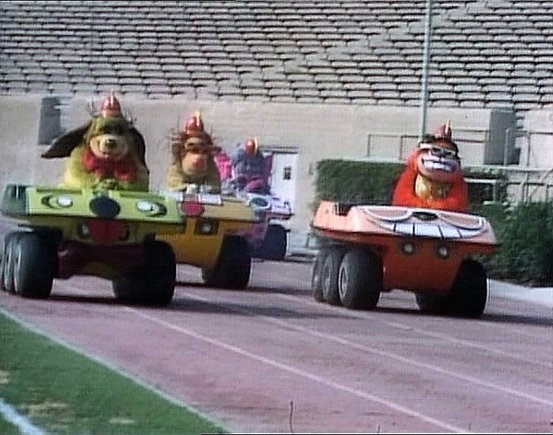 Six wheeled cars - Page 2 - General Gassing - PistonHeads - The image captures a lively scene from a parade or a race. It features three cartoonish trucks, each decorated and manned by people dressed in colorful costumes that resemble animals. These individuals appear to be participating in the event, riding on top of their respective vehicles which are styled as race cars. They're driving on what seems like a track, as indicated by the markings visible behind them. The setting is outdoors under clear skies, and there are spectators present, suggesting a public event with an audience.