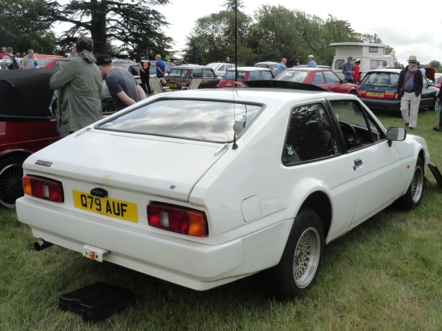 RE: Festival of the Unexceptional | PH Gallery - Page 8 - General Gassing - PistonHeads - The image features a classic white sports car parked in an outdoor setting. It appears to be on display, likely at a car show or similar event. There are people milling about the area, suggesting that it's a public event of some sort. In the background, there's a grassy area and what looks like a small structure or perhaps a tree with dark foliage. The car itself is quite striking, with its white body contrasting against the green surroundings. It's positioned at an angle to the camera, giving us a clear view of its sleek design and distinctive features.