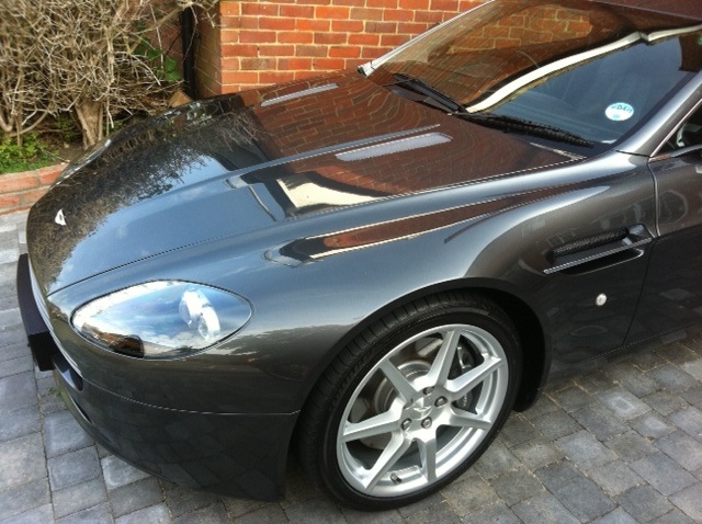 Clay bar treatment - Page 1 - Aston Martin - PistonHeads - This image shows a close-up view of the front half of a gray luxury car parked on a brick driveway. The car's sleek design, highlighted by a large, accent-lit grille, is the focal point. The grille is capped with what appears to be a reflective, metallic material. The headlight is modern, with an angular design, and is adorned with a small, emblem-like triangle at its center. The tire is neatly tucked under the car's aerodynamic wheel arch, and the road is blurred in the background, suggesting a focus on the vehicle itself. The car's side mirrors are also visible, confirming the view of the front half. The brick driveway provides a textured and rustic backdrop to the car's high-end appearance.