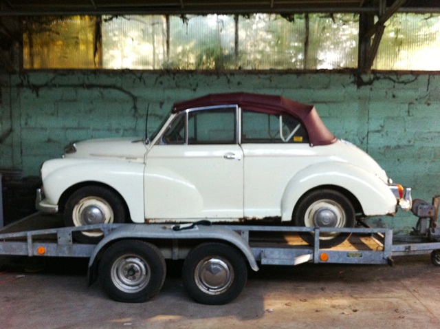 A truck parked on the side of a road - Pistonheads - The image shows a vintage white car with a brown convertible top resting on a trailer. This trailer is equipped with triangular safety flags at each end for safety purposes. The vehicle is likely being towed, possibly for restoration, as it is described as old and being prepared for a trip. The setting appears to be on a concrete surface, and the background features a structure with corrugated metal and foliage, suggesting an outdoor or semi-outdoor location.