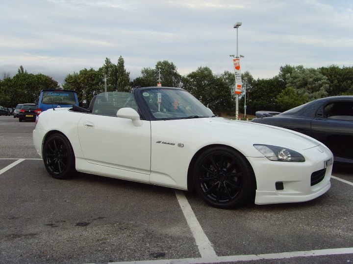 Pistonheads - The image depicts a white sports car parked in a parking lot. The car is oriented at a slight angle to the camera, showing the front and side of the vehicle. A person with a backpack is visible in the front seat, suggesting that the car might be used for travel or daily commuting. Beyond the white sports car, there are other cars parked in the lot, and a sign with the word "info" on it along with some other indistinct signs. The sky is overcast, providing a neutral backdrop.