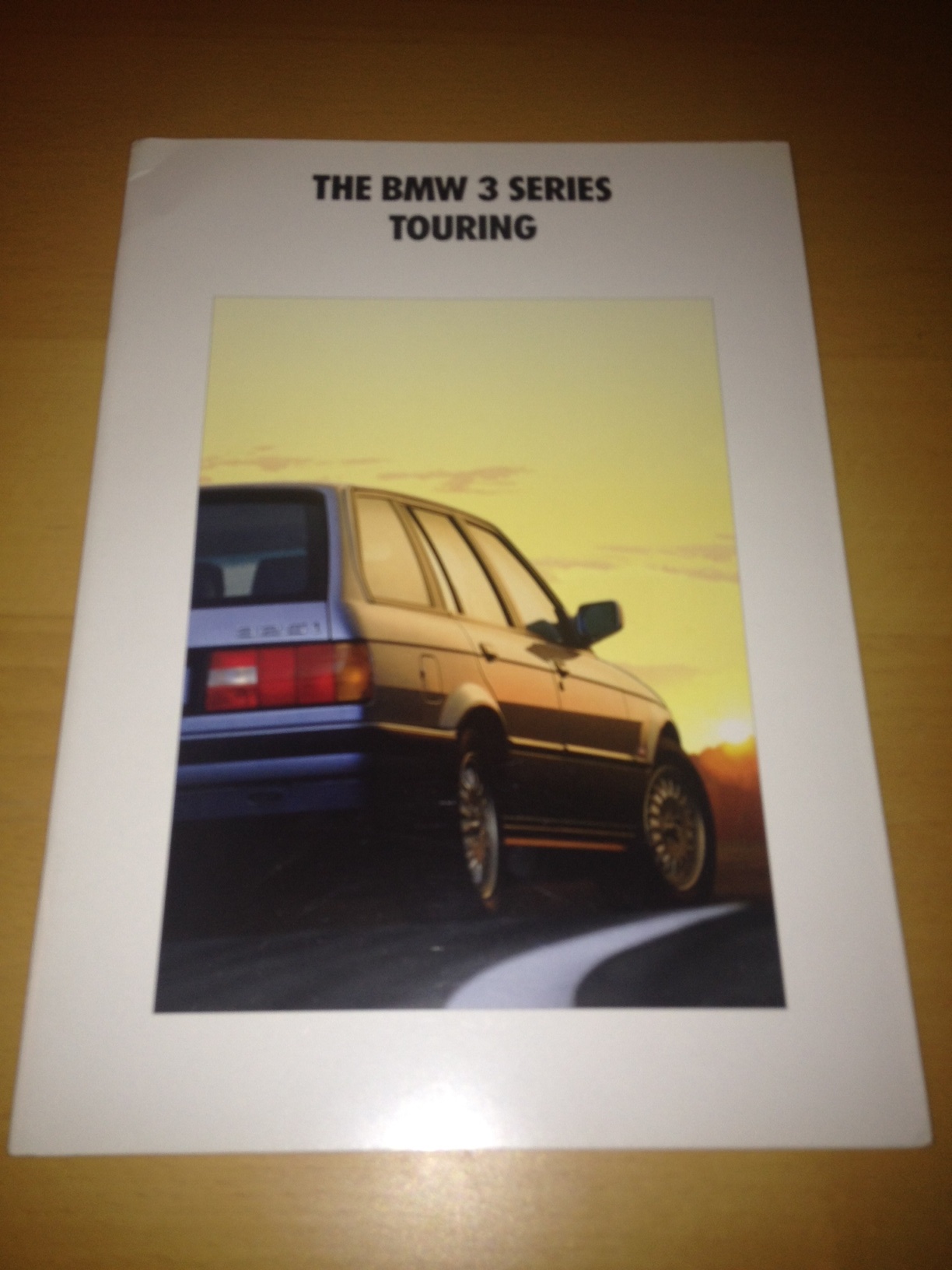 RE: Shed of the Week: BMW 3 Series Touring (E30) - Page 1 - General Gassing - PistonHeads - The image features a brochure about the BMW 3 Series Touring. The BMW logo is prominently displayed in the top left corner. The back of the brochure features a picture of a classic BMW 3 Series car, likely intended to showcase the brand's heritage and appeal. The text urges viewers to learn more about BMW and its offerings. The brochure is placed on a wooden surface, suggesting it might be in front of a potential buyer or enthusiast.