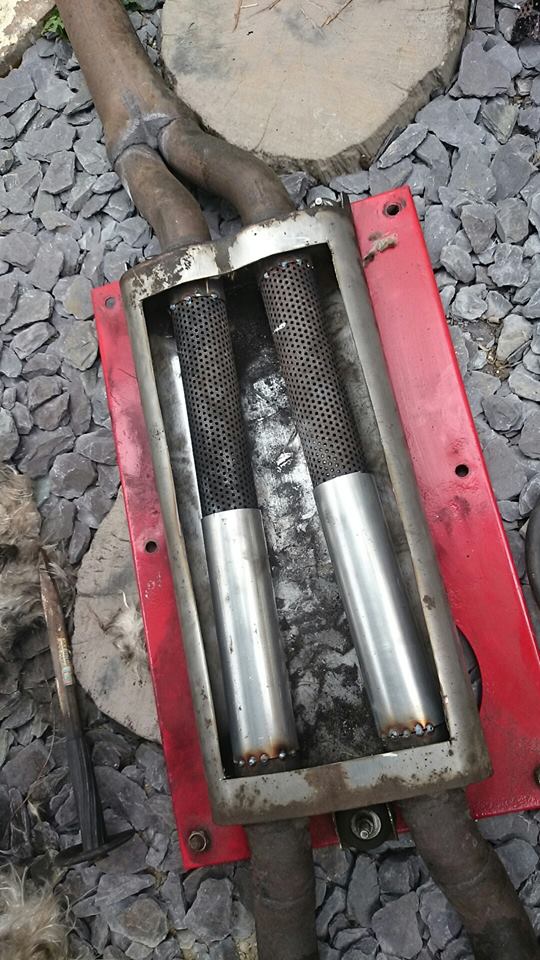 A pair of rusty scissors sitting on the ground - Pistonheads - The image shows a rusted, metallic object composed of several cylindrical sections mounted on a red base. The cylindrical parts appear to be stained or rusted, and there are two visible central components that stand upright. The surrounding area includes what looks like rocky terrain pebbling the floor. A smaller, black object is partially visible in the lower left corner of the photo, though its purpose is unclear. The object is photographed with an angle that emphasizes the side view of the metallic structure.