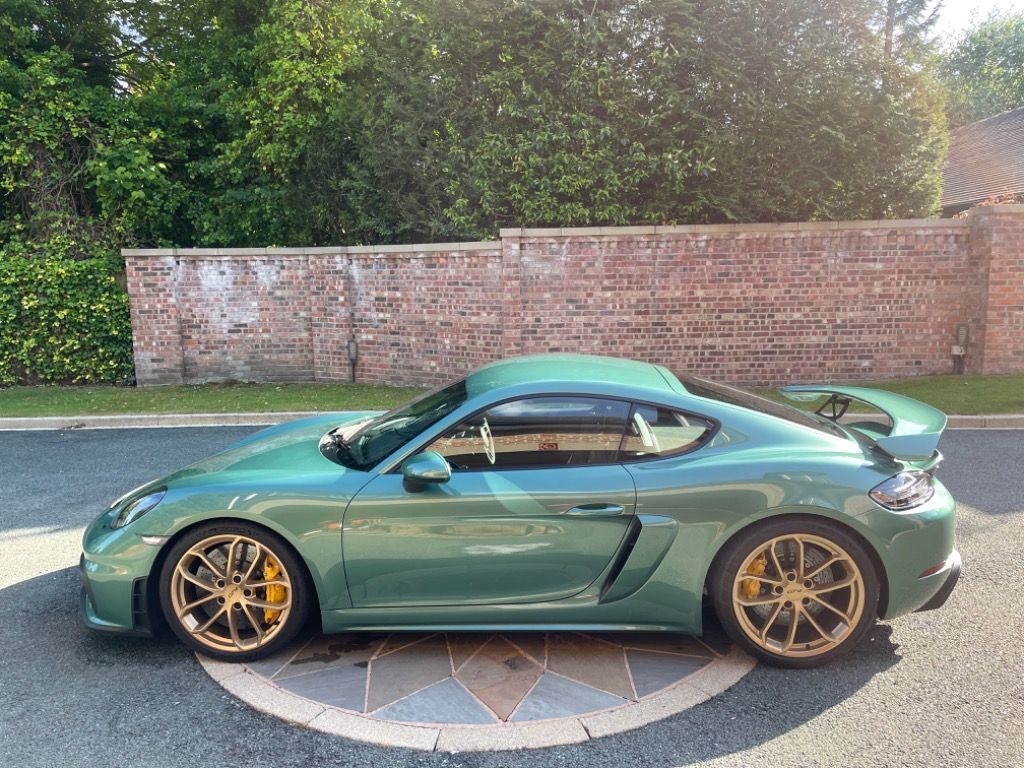 718 GT4 UK Allocations  - Page 101 - Boxster/Cayman - PistonHeads UK - This is a photo of a green sports car parked on the street. The vehicle appears to be in good condition and is likely a high-end model, as indicated by the features like the low and wide stance, wide arches, and prominent spoilers. It's positioned on what looks like a custom roundabout or island. There are no visible people in the image. The background shows a residential area with brick walls and greenery, suggesting a suburban setting. The sky is overcast, indicating either dawn or dusk.