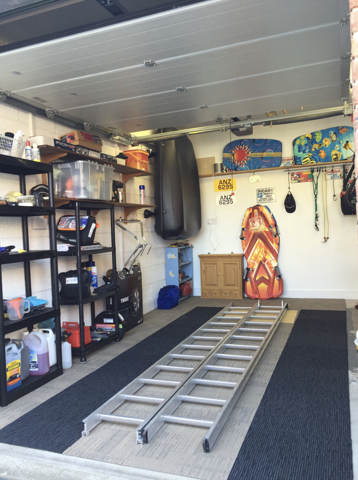 A room filled with lots of wooden furniture - This image shows the interior of a garage, which is equipped with various items. On the left side of the photo, there's a black shelf containing what appears to be household cleaning supplies and some tools. In the center of the image, a set of steps is leaning against the wall, suggesting recent use or storage. To the right, there are personal belongings including a surfboard mounted on the wall, a boogie board placed upright, and a black gym bag with yellow writing resting against the wall. The flooring of the garage appears to be concrete, and the walls are painted white.