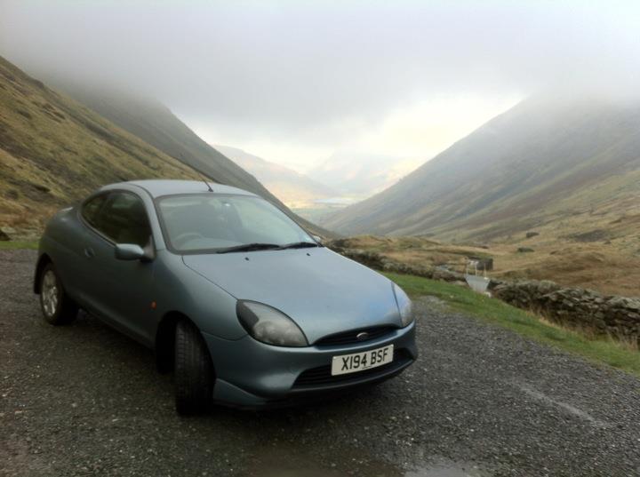 Help identify this Lake District location? - Page 1 - General Gassing - PistonHeads - This image features a dark-colored Ford Fiesta car parked on a gravel surface. It's set against the backdrop of a dramatic mountainous landscape. A short road or path leads to the car, hinting at a remote area. The weather appears to be somewhat gloomy with cloud cover, and the lighting suggests an overcast day. The presence of a license plate indicates the car is on a public road.