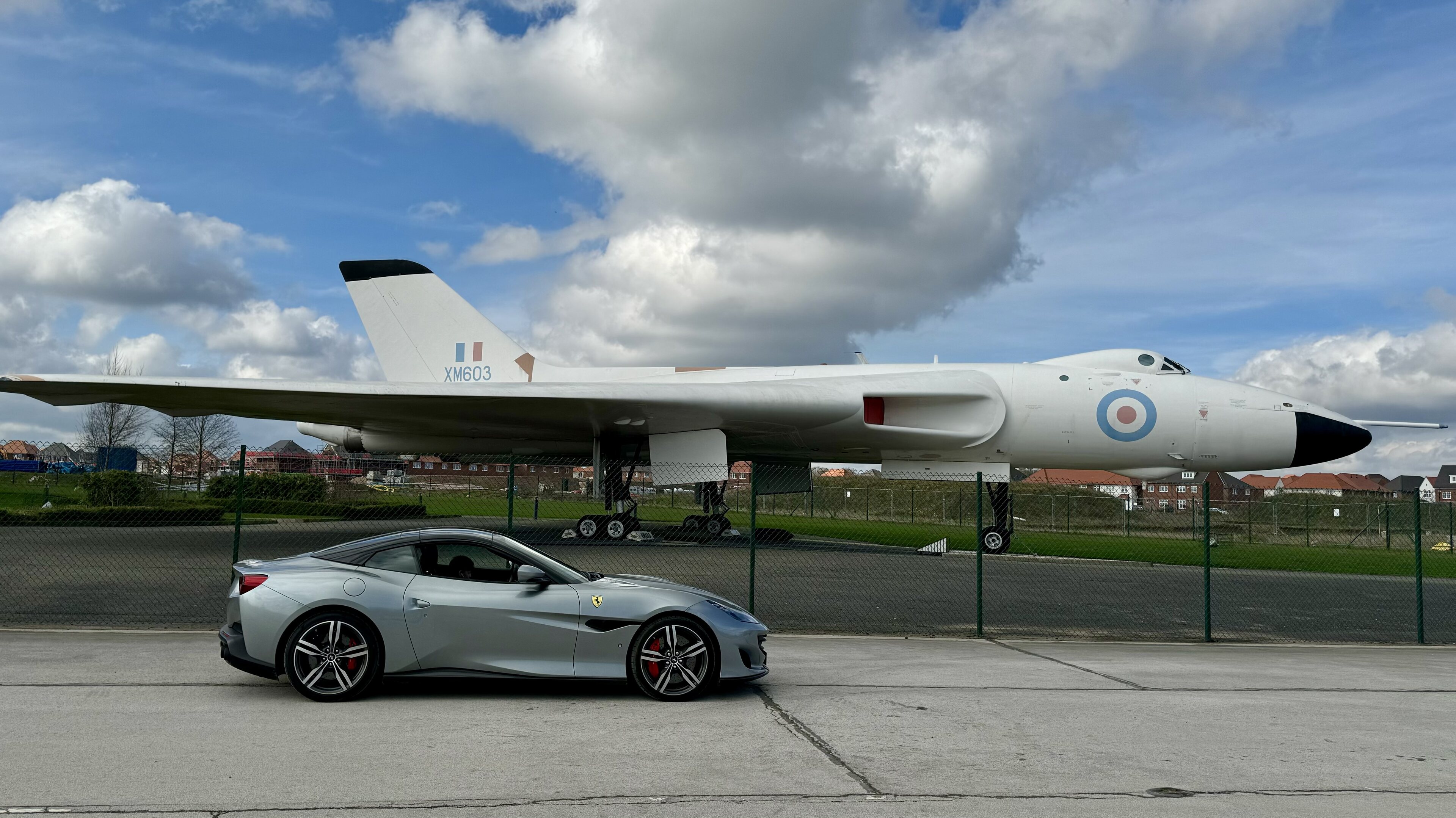 Vulcan bomber info  - Page 2 - Boats, Planes & Trains - PistonHeads UK - The image depicts a scene at an airfield. In the foreground, there's a silver car parked on the tarmac. Behind it, a large military aircraft is prominently displayed. The jet has a camouflage pattern and bears the markings of a particular military service. The sky is clear with a few scattered clouds, indicating good weather conditions. There are also some fences and trees visible in the background, suggesting the presence of other facilities or vegetation around the airfield.