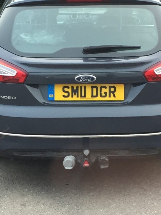 What crappy personalised plates have you seen recently? - Page 488 - General Gassing - PistonHeads - The image shows the back of a dark-colored car with a visible license plate. The license plate is rectangular with a yellow background and black text, reading "SMU DGR". Behind the license plate, there is a towing eye, commonly known as a spare tire mount, with a circular silver cover. The car also features two red taillights and a clear, glossy rear window behind which there are no visible items or objects. The Ford emblem is on the trunk, indicating the make of the vehicle. The image appears to have been taken during daylight, and the focus is on the registration details of the vehicle. The style of the image is a real-life photograph with a clear, direct shot of the car's rear detail.